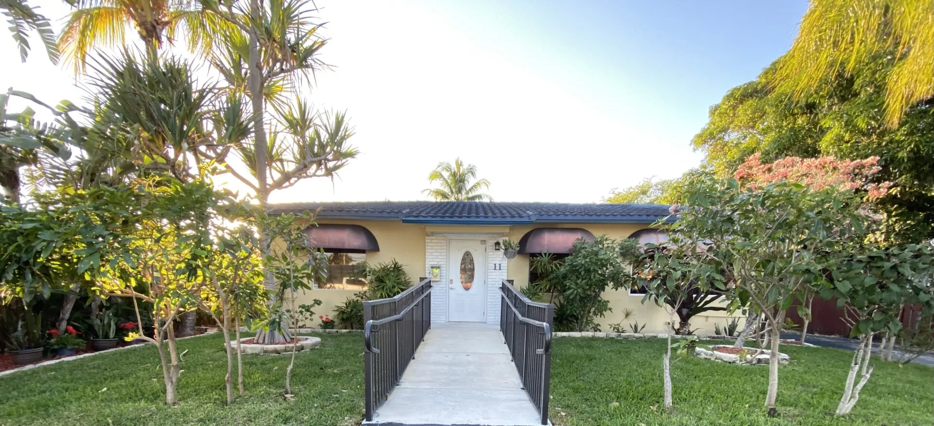 Single-story tan house with dark roof, white front door, and metal railings flanking a concrete pathway through manicured lawn and tropical landscaping