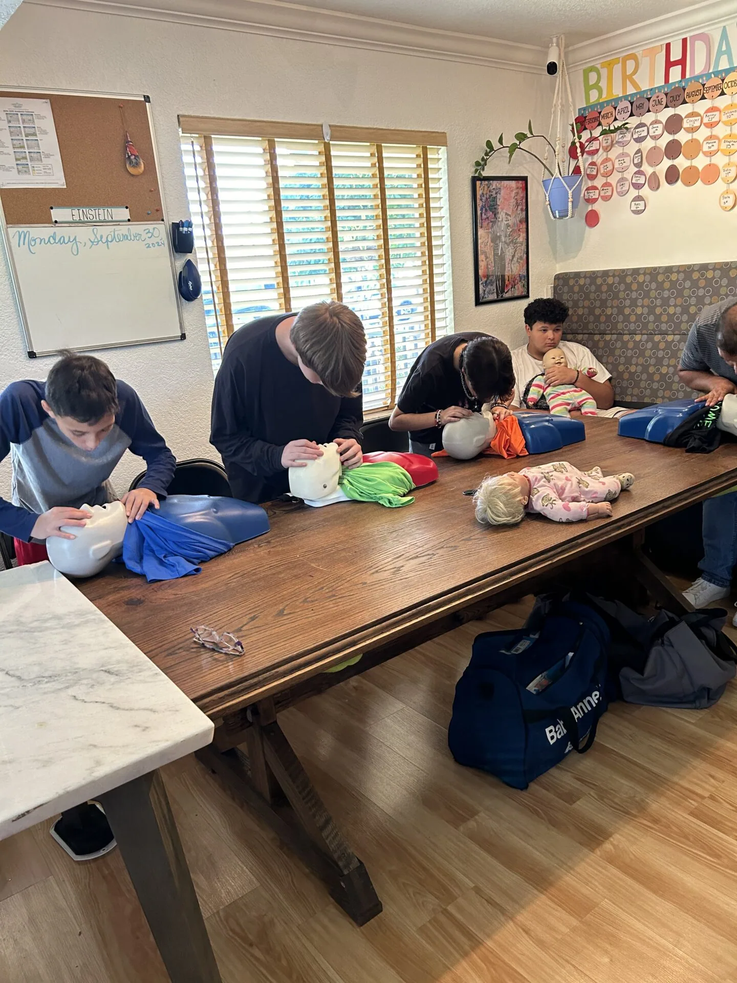 Students seated at a wooden table in a classroom, working with dolls and fabric materials during a lesson