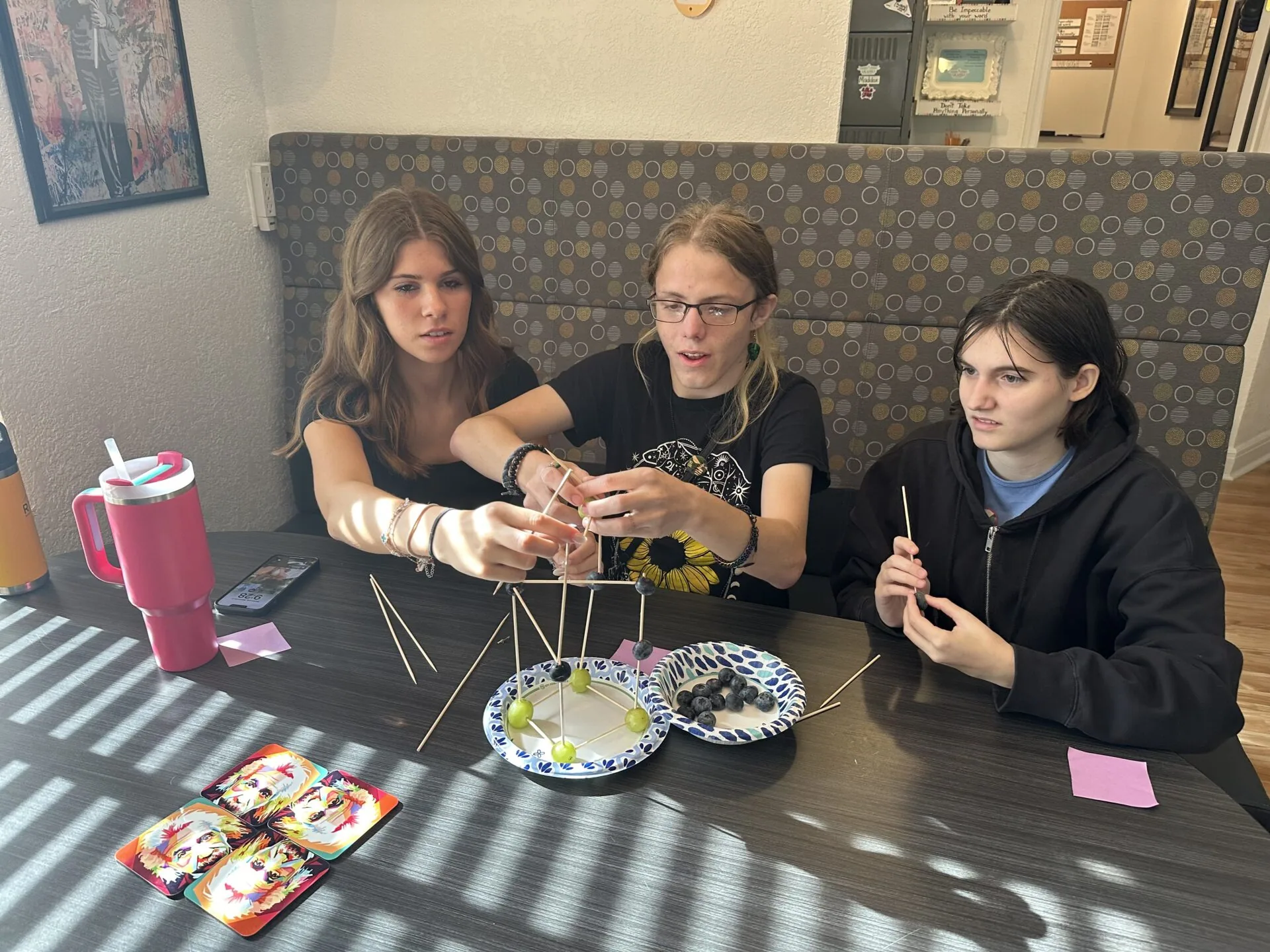 Three young women sitting on a couch at a table with craft supplies and food items, appearing to work on a hanging mobile or wire structure project