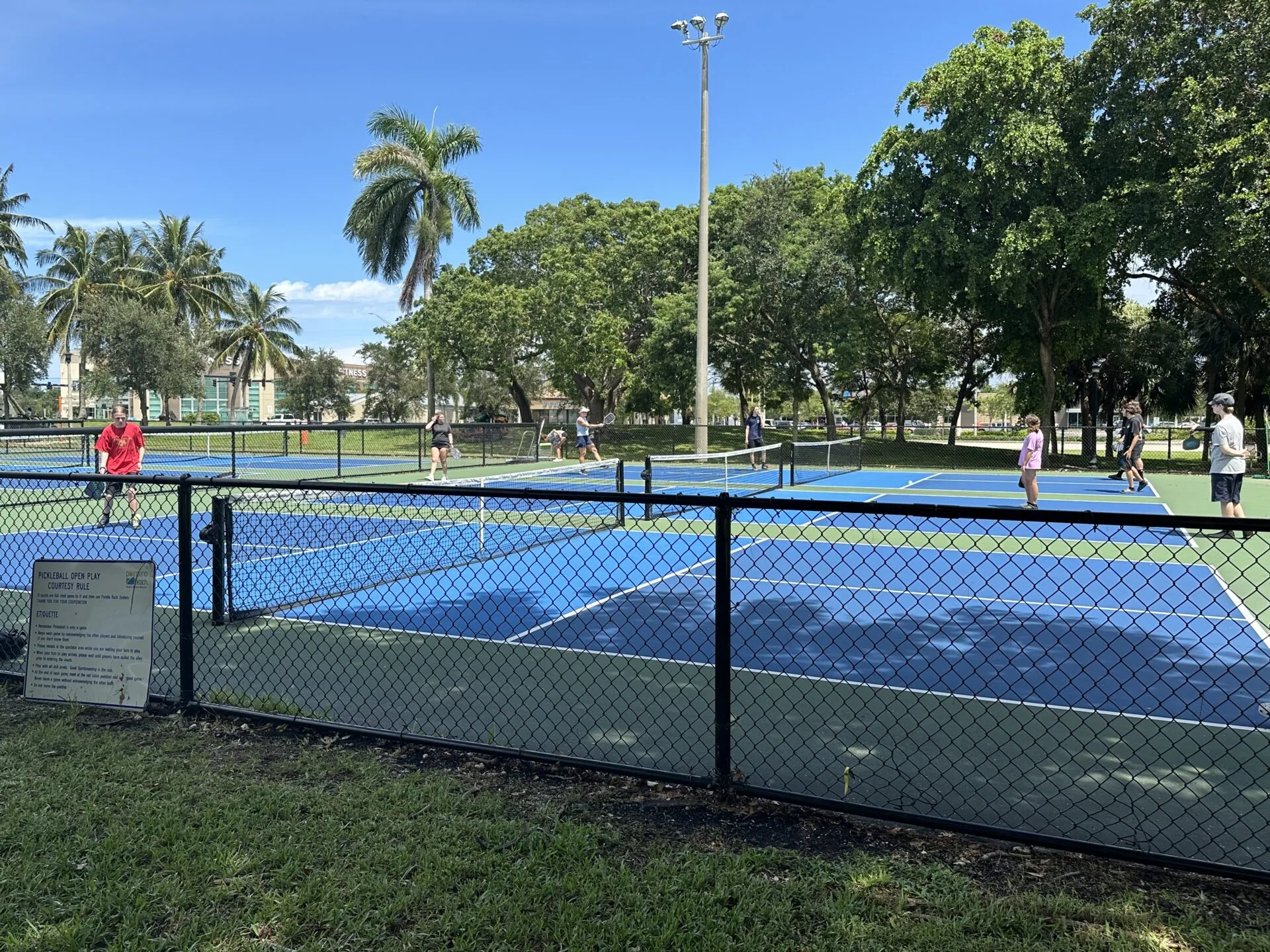 Public park with blue tennis courts surrounded by chain-link fencing, palm trees, and recreational areas on a clear day