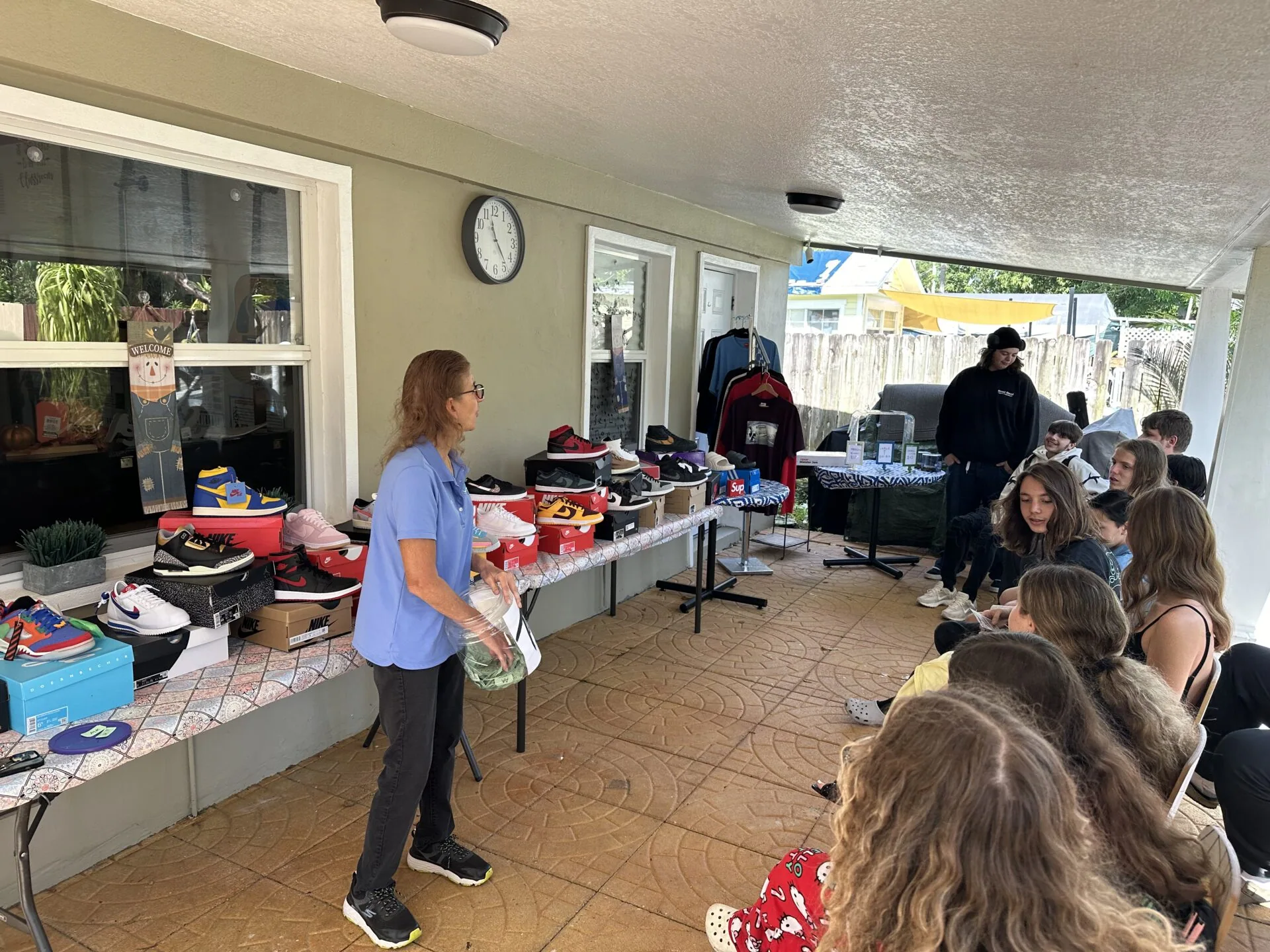 Woman in blue shirt standing in covered outdoor space with tables of shoes and children browsing items during what appears to be a community event or sale