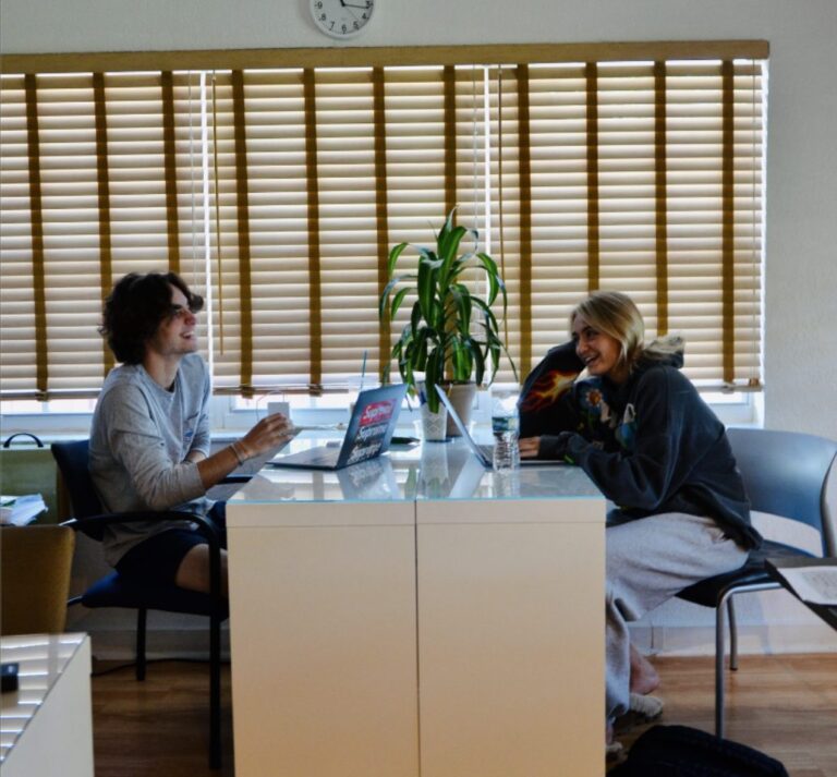 A tutor and a student work across a white desk in a bright room with shutters
